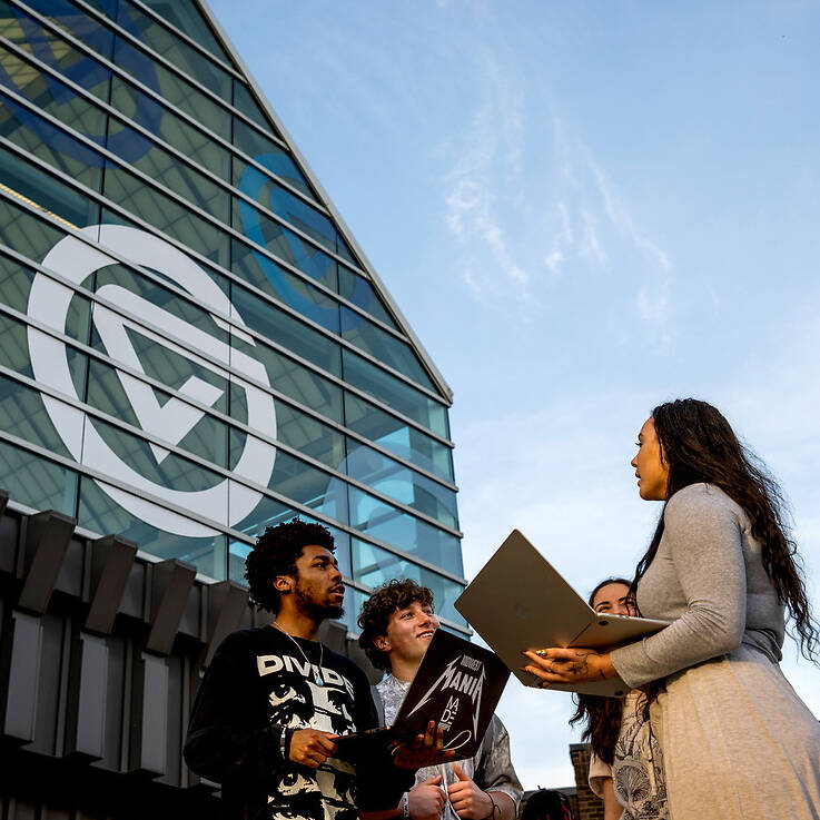 A group of individuals stands outdoors in front of a modern glass building with a large circular logo, engaging in conversation under a clear blue sky.